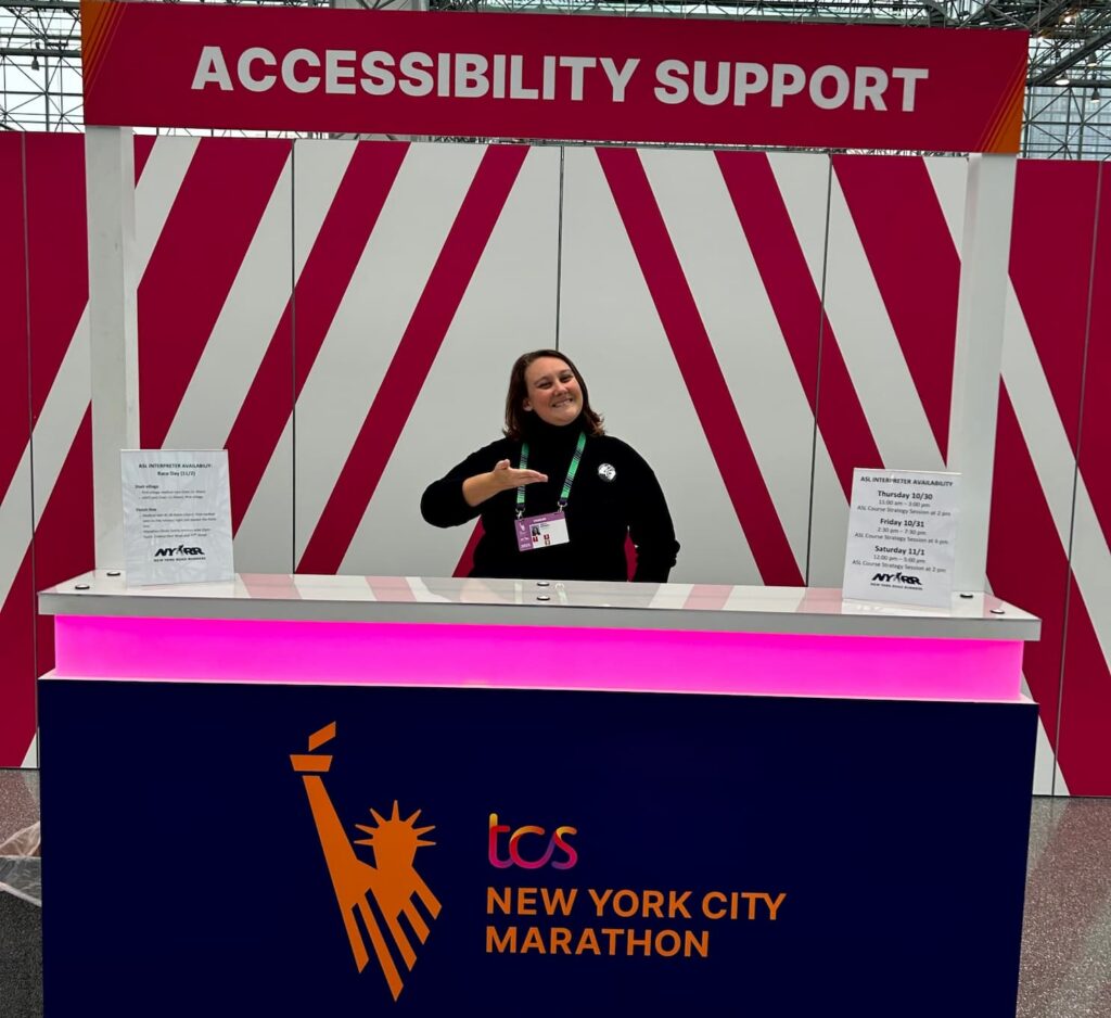 An ICS team member signs from behind the TCS New York City Marathon Accessibility Support booth, with printed ASL interpreter availability schedules posted on the counter at either side.