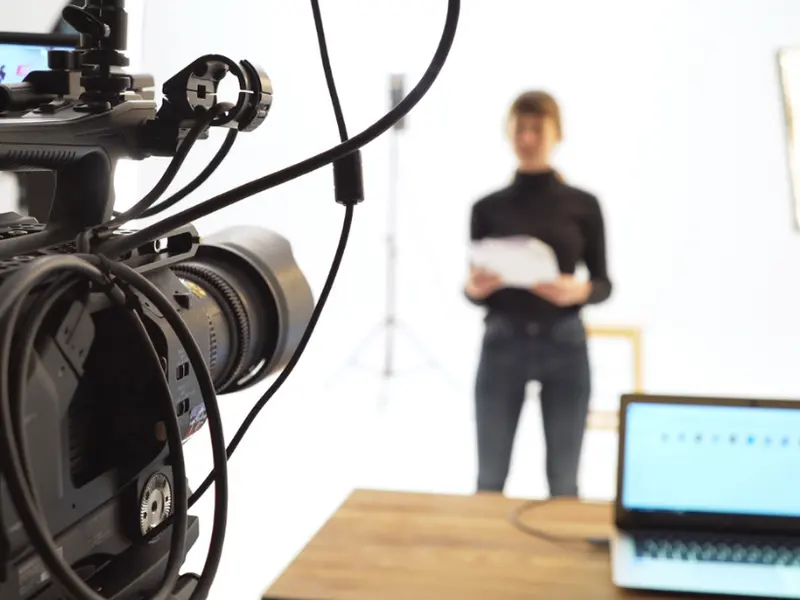 A video camera in a bright studio filming a presenter holding a script