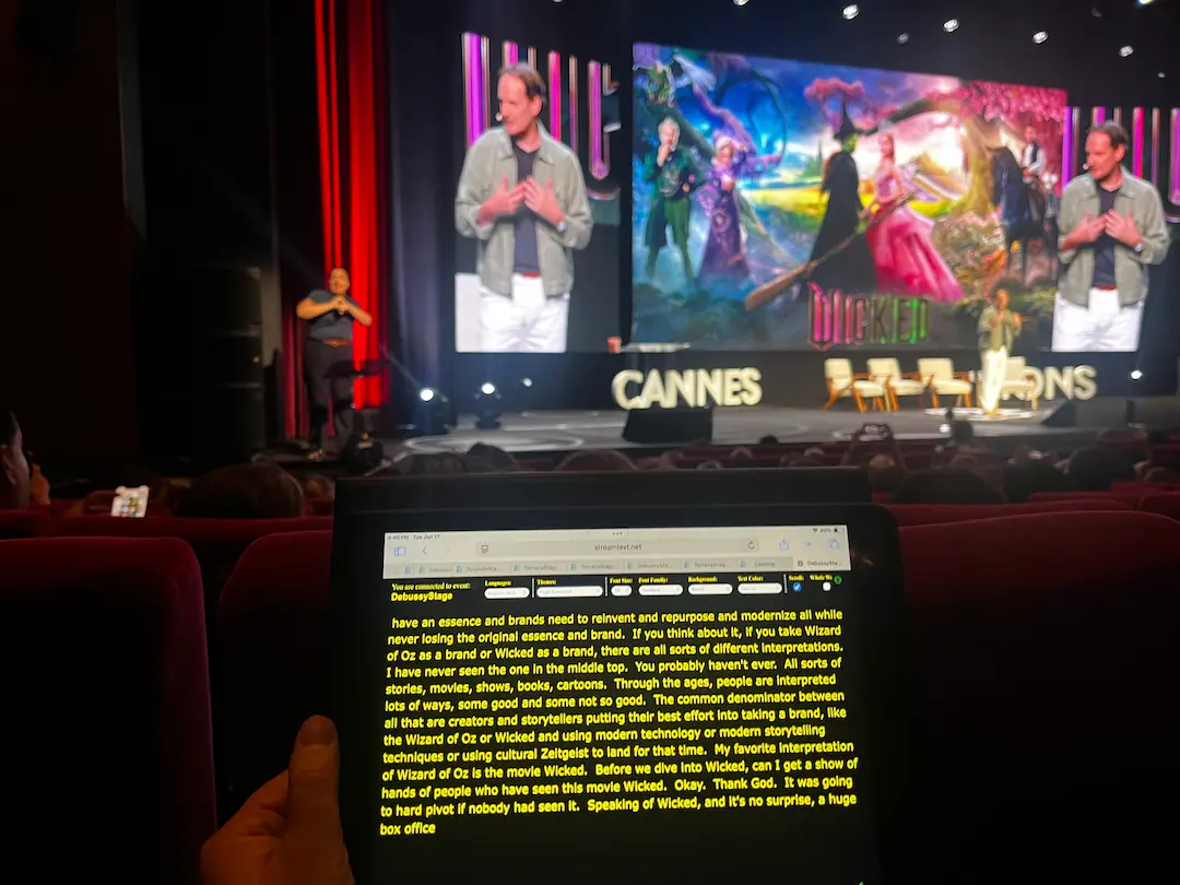 View from the audience inside a theater. On stage, a presenter is speaking during a session about the movie Wicked. An ASL interpreter stands to the far left of the presenter. In the foreground, an audience member follows along using CART captions on their personal iPad.
