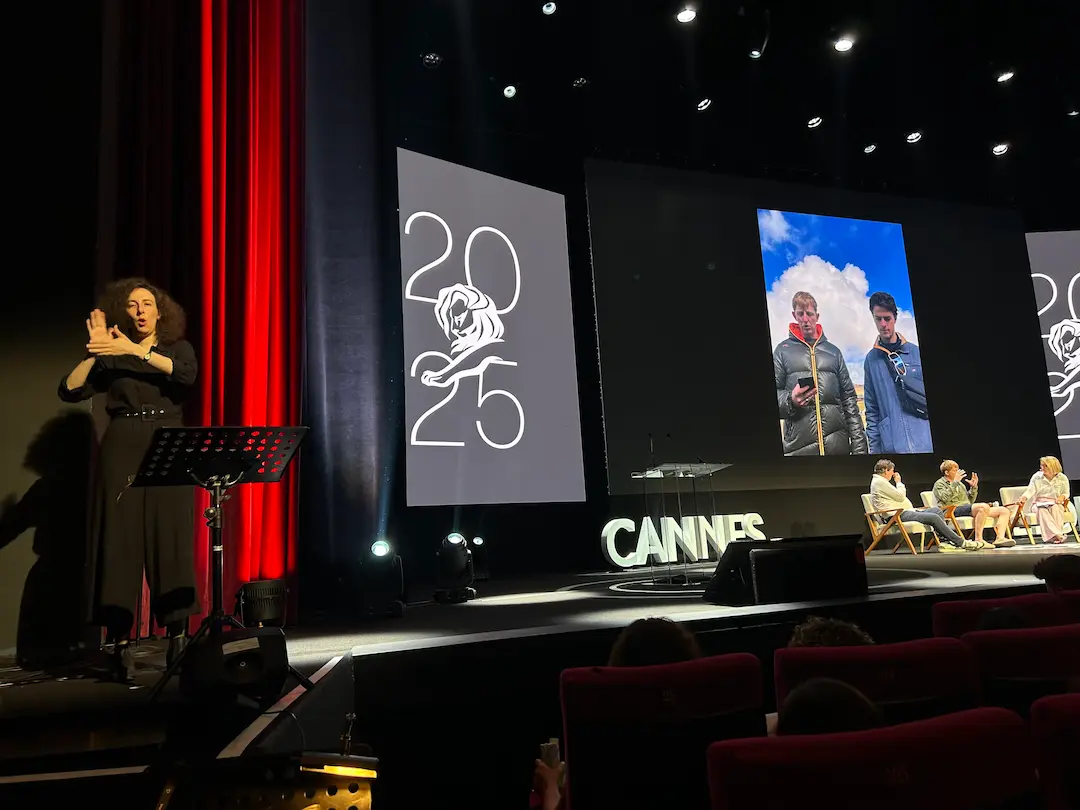 When the lights dim, access fades, image one of two: an interpreter stands stage left on the Cannes Lions main stage, clearly visible and well-lit while a panel session runs behind her.