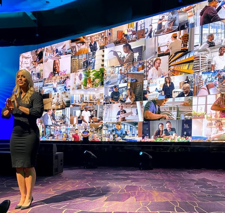 An ASL interpreter signs at the edge of the stage in front of a wide LED wall filled with a grid of customer photographs from the keynote presentation.