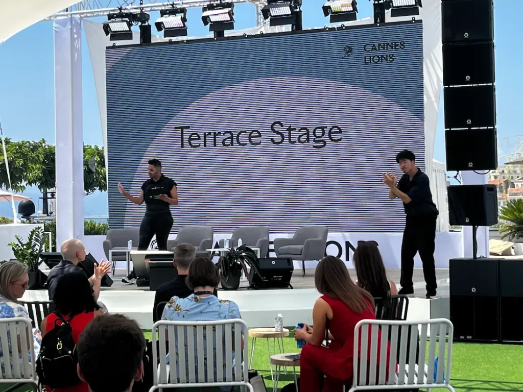 An outdoor stage with a large projected screen. The speaker stands on the main platform, while an ASL interpreter is visible on a smaller platform to the right.
