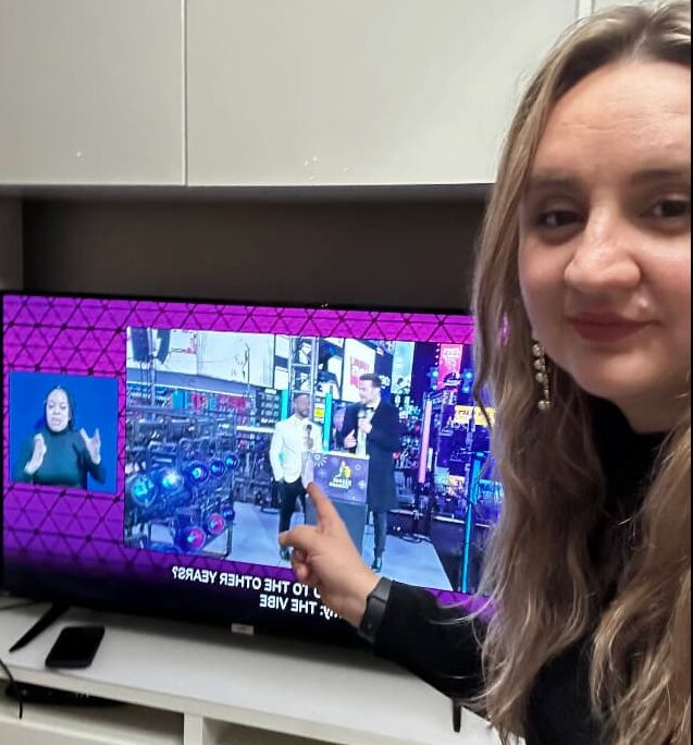 A viewer at home smiles at the camera while pointing back at her television, which is showing the Times Square broadcast with the ASL interpreter panel in the corner.