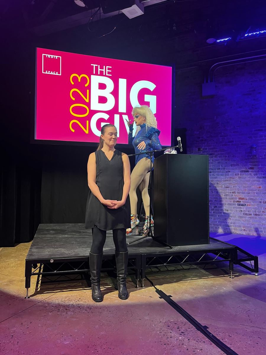 An ASL interpreter is standing in front of a black stage and a large tv screen. The screen has a pink background with yellow and white lettering reading "2023 the Big Give."