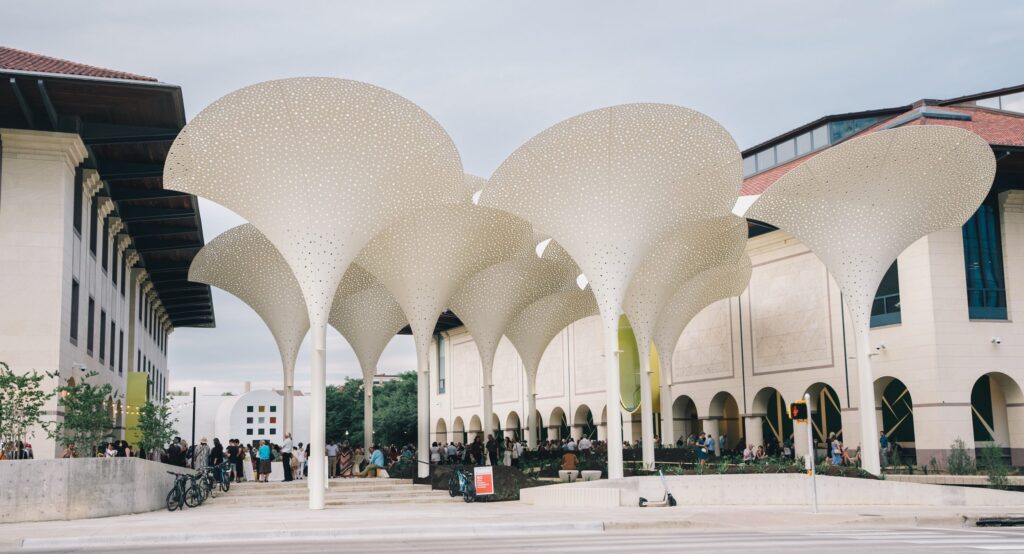 A crowd standing and talking on a museum forecourt under a grove of tall white perforated canopies shaped like petals, an arcaded stone building and parked bicycles behind them.