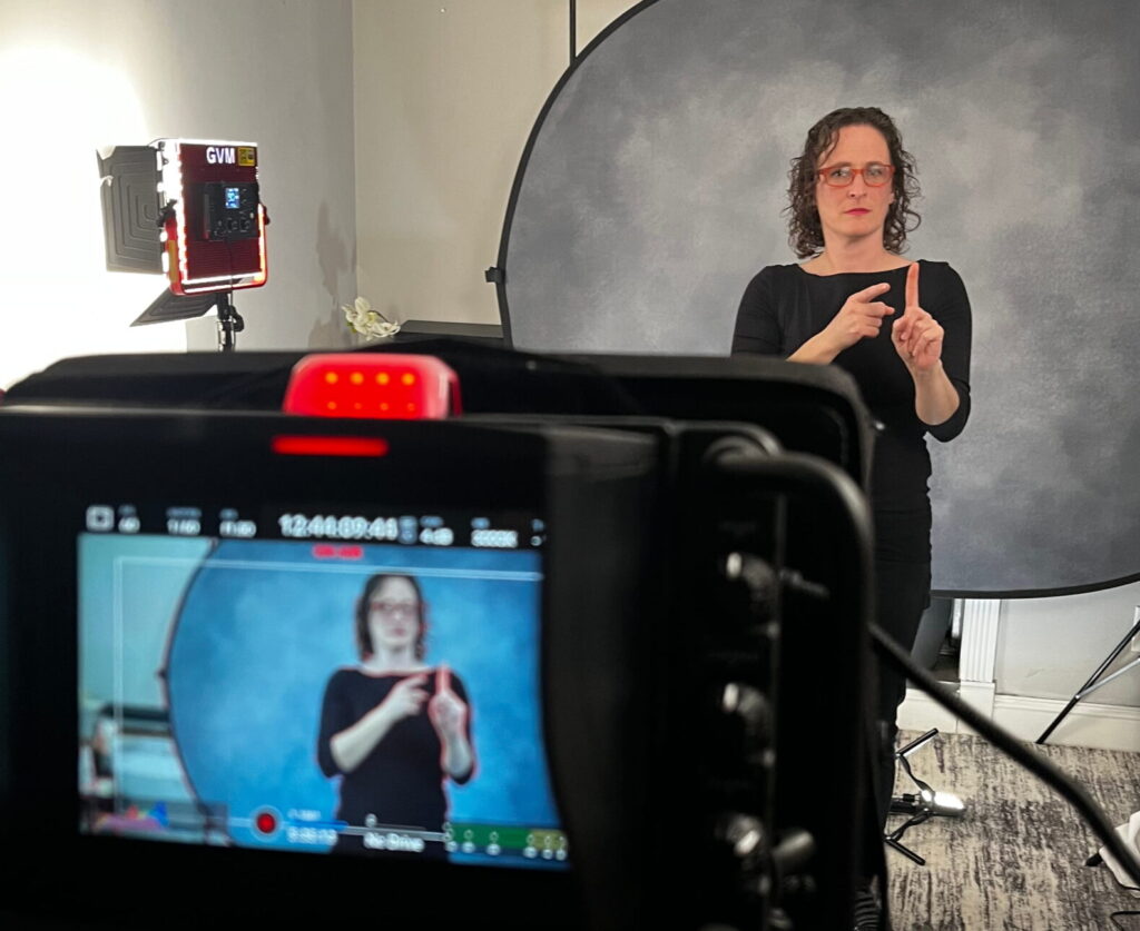 The studio setup during a take: the Deaf interpreter signs in front of the grey backdrop under an LED panel, framed on the camera monitor in the foreground with the recording timecode running.