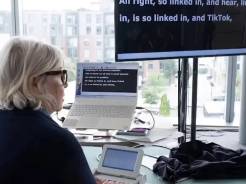 A captioner typing on a stenotype machine while facing the large screen where live captions appear