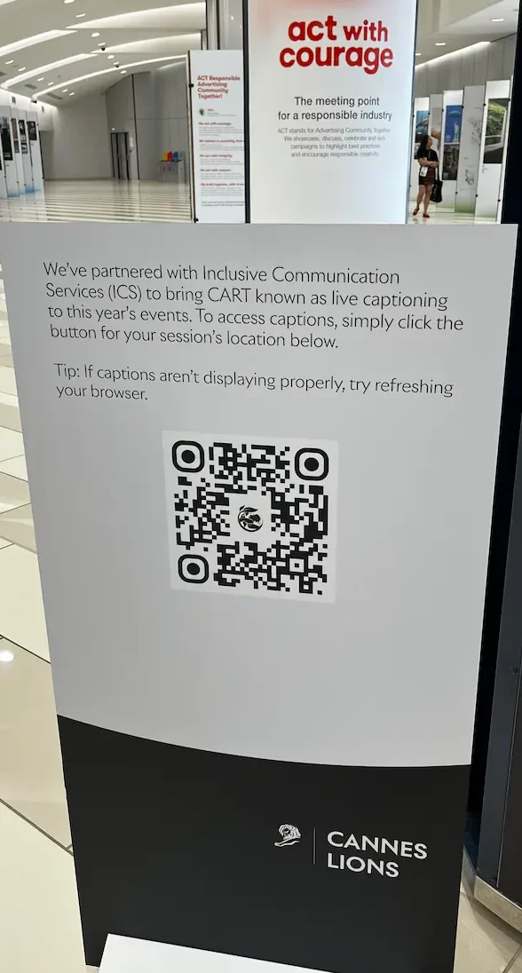 A printed poster in a festival corridor reading 'We've partnered with Inclusive Communication Services (ICS) to bring CART known as live captioning to this year's events. To access captions, simply click the button for your session's location below,' above a QR code.