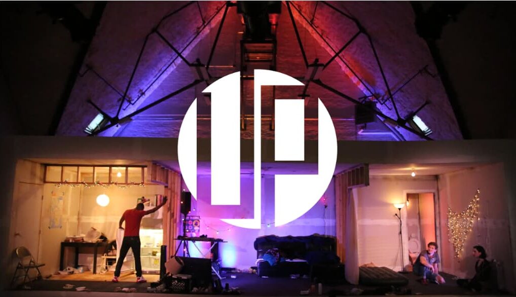 The UP Theater Company mark over a production still: a full stage set built inside the church nave, a lit apartment interior on the left and a bedroom on the right, actors in place under coloured lighting rigged from the vaulted ceiling.