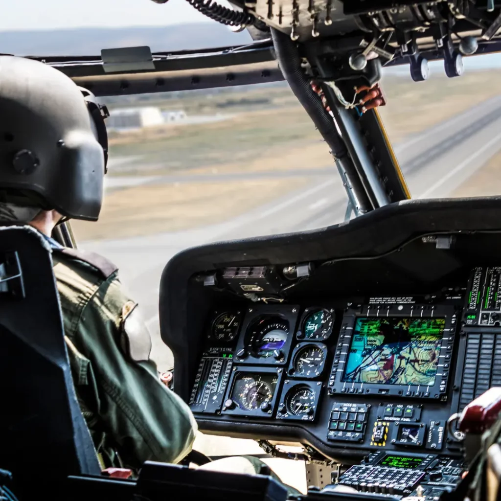 The illustration the source page used for this section: a helmeted pilot at the controls of a helicopter cockpit, instrument dials and a moving-map display filling the panel, with a runway visible through the windscreen.