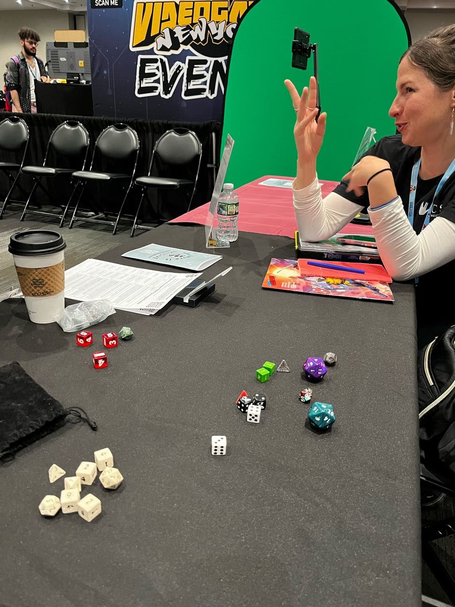 Rebecca is seated at a long table with a black tablecloth. The table has game dice on it. Rebecca is signing in ASL. In the background is a sign that reads "Video Game New York Events.”