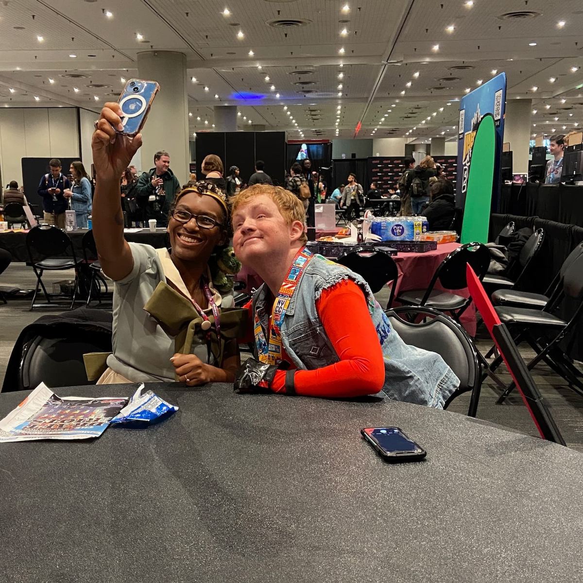 Two people are sitting at a table smiling taking a selfie together. The person on the left is wearing a light blue shirt with a large green bow, holding the phone. The person on the right is wearing a red shirt with a denim vest and black gloves.