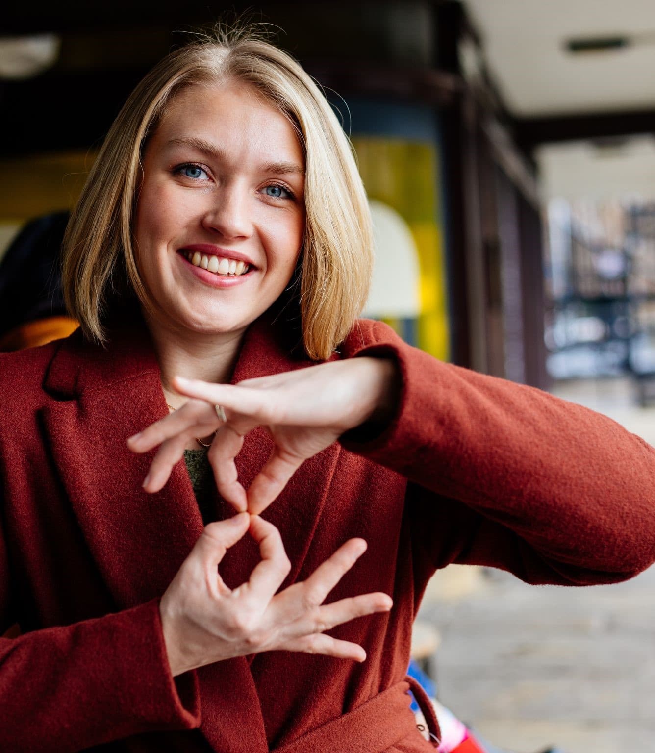 A smiling woman with a blonde bob, wearing a rust-colored coat, stands on a city street with both hands raised near her chest, thumb and fingertips of each hand pinched together.