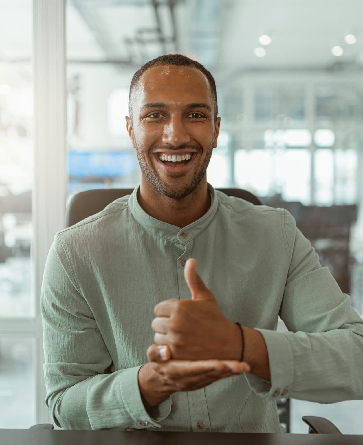 A smiling man in a light sage-green shirt, at a desk in an office with large windows; one hand is closed with the thumb pointing up, resting on the open palm of his other hand.