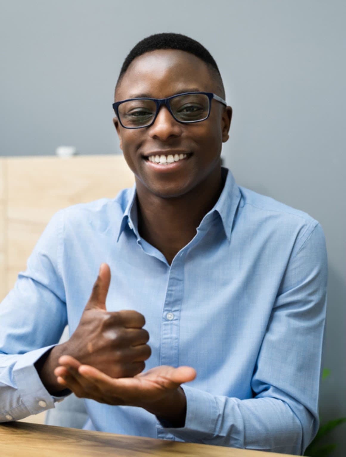 A smiling man with glasses, wearing a light blue shirt, seated near a wooden cabinet; one hand is closed with the thumb pointing up, resting on the open palm of his other hand.