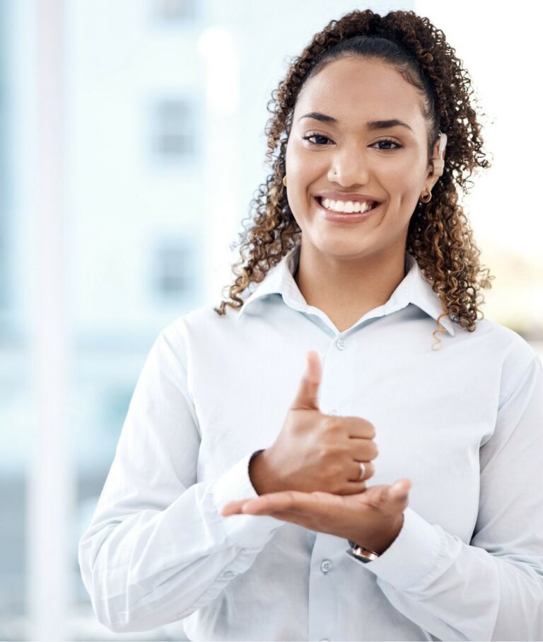 A smiling woman with curly hair, wearing a white collared shirt, in a bright office; one hand is closed with the thumb pointing up, resting on the open, upturned palm of her other hand.