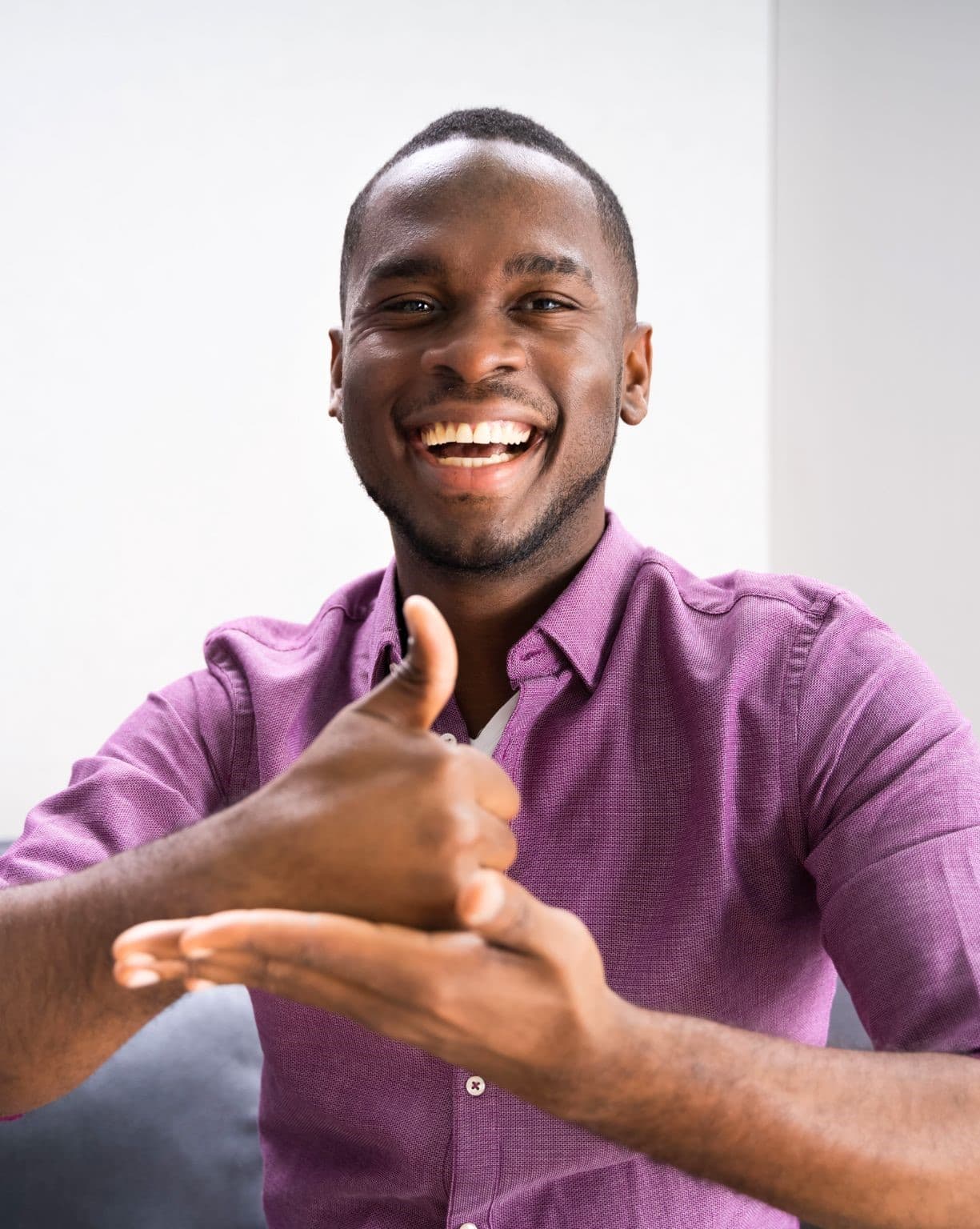 A smiling man in a purple button-down shirt, photographed against a plain light background; one hand is closed with the thumb pointing up, resting on the open palm of his other hand.