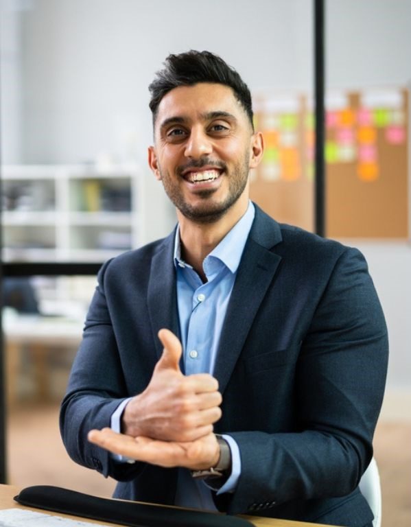 A smiling man in a navy suit jacket at a desk, with a corkboard of sticky notes behind him; one hand is closed with the thumb pointing up, resting on the open palm of his other hand.