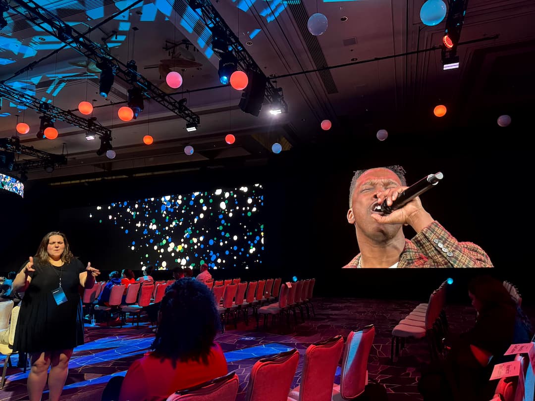 An ASL interpreter signs from the aisle at the side of the ballroom during a musical performance, with the performer singing into a microphone on the screen beside her and coloured globe lights hanging from the lighting rig overhead.