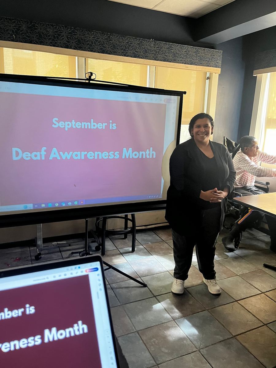 A woman is smiling and giving a presentation. She is in front of a projection screen with red with white letters that reads “September is Deaf Awareness Month.”