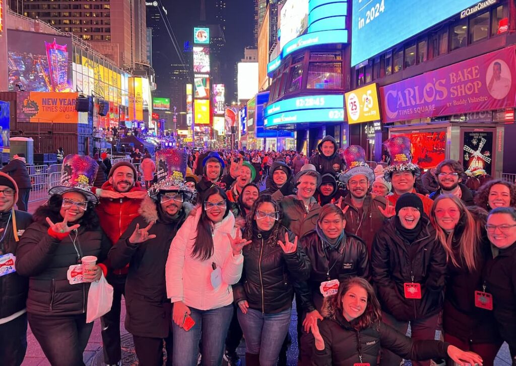 A large group of Deaf attendees packed together on the street in Times Square before midnight, many wearing 2024 novelty glasses and party hats and several signing to the camera.