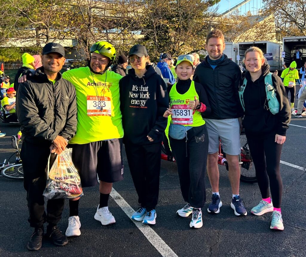 Six runners and supporters pose together in the staging area before the race, two of them wearing Achilles International singlets over TCS New York City Marathon bibs.