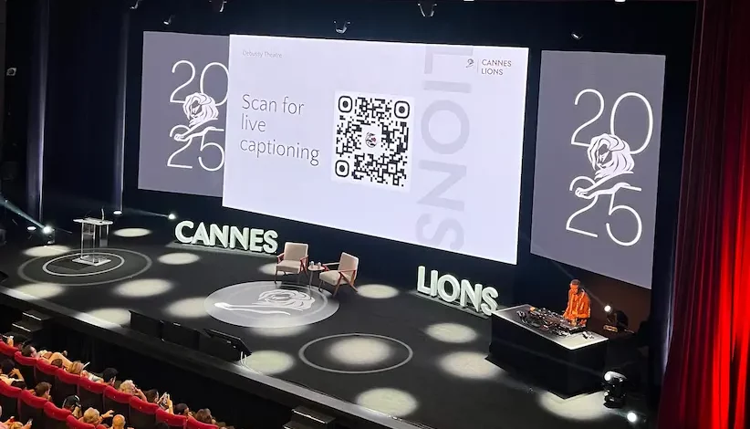 The Debussy Theatre main stage at Cannes Lions 2025, with a slide on the large screen reading 'Scan for live captioning' beside a QR code, above an empty two-chair set.