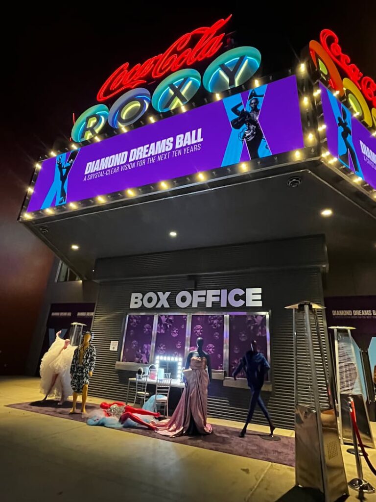The Coca-Cola Roxy marquee lit at night, carrying a Diamond Dreams Ball banner that reads 'A crystal-clear vision for the next ten years' above the venue box office, with mannequins in couture gowns arranged on the sidewalk below.