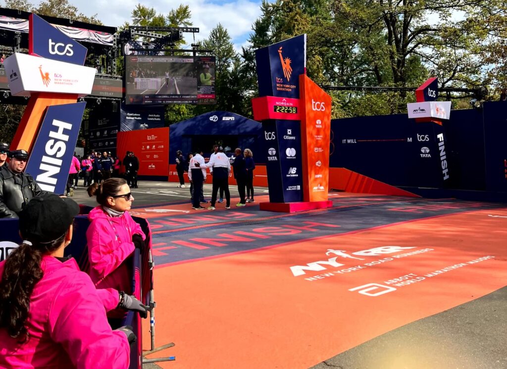 The TCS New York City Marathon finish line in Central Park, with the race clock reading 2:22:58 above the orange finish carpet and staff in pink jackets watching from the stands.