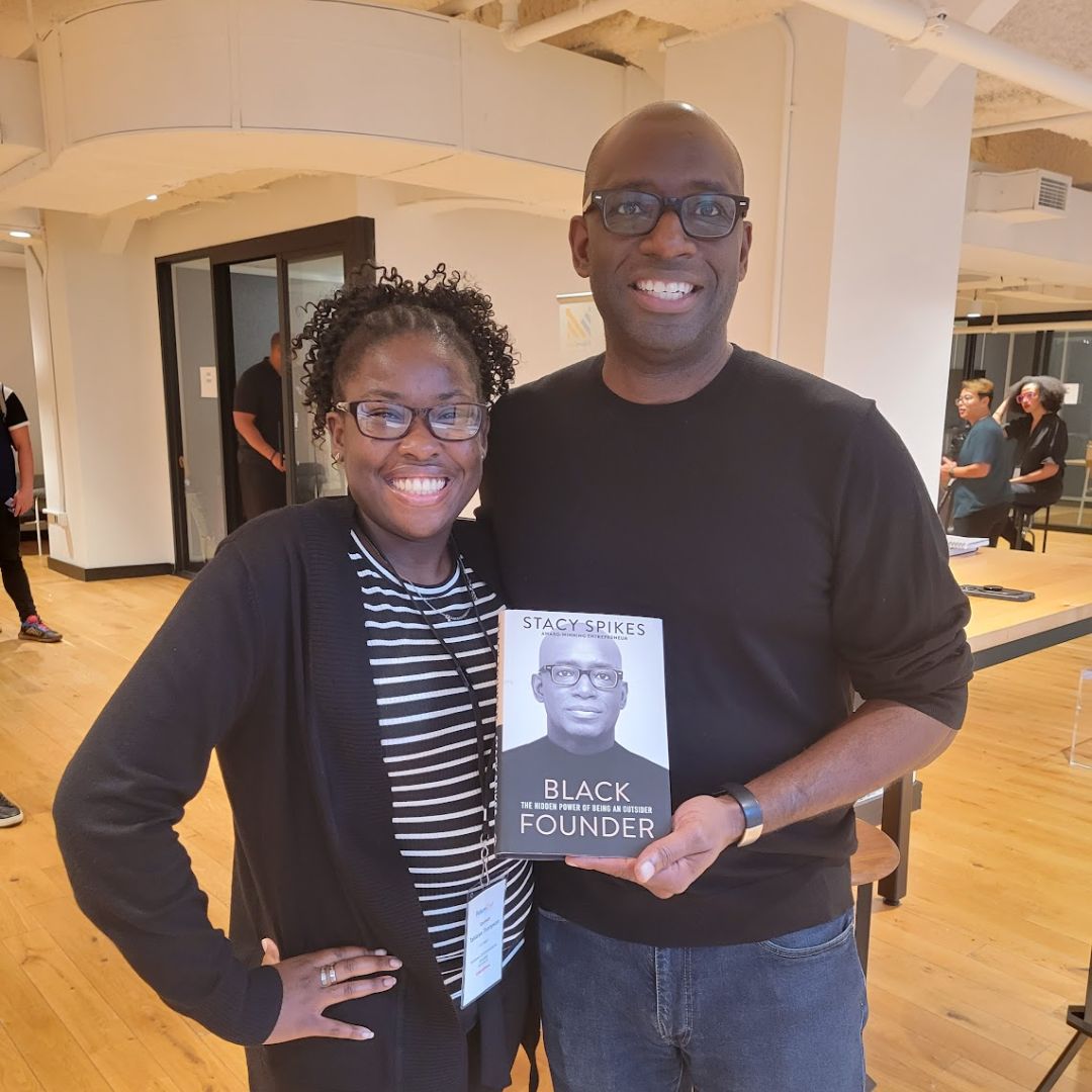Two people posing for a picture in a well-lit room. The man is holding the book with his picture on it. The book is called “Black Founder: The Hidden Power of Being an Outsider" by Stacy Spikes. An ICS intern is standing next to him, and both of them are smiling.