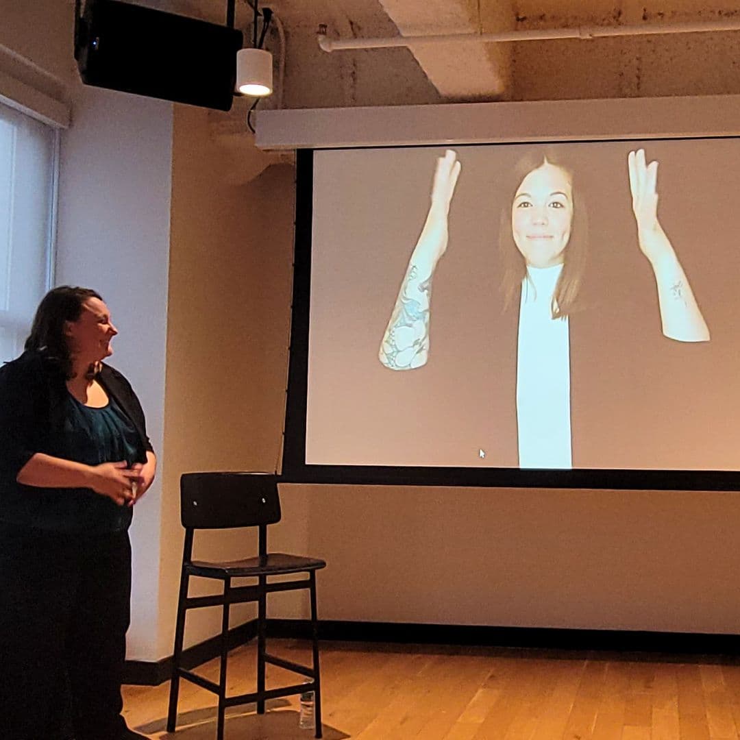Shelby is giving a presentation in a room. On the projection screen is Kaycee signing. In front of the projector is an empty black chair and a woman is facing the screen.