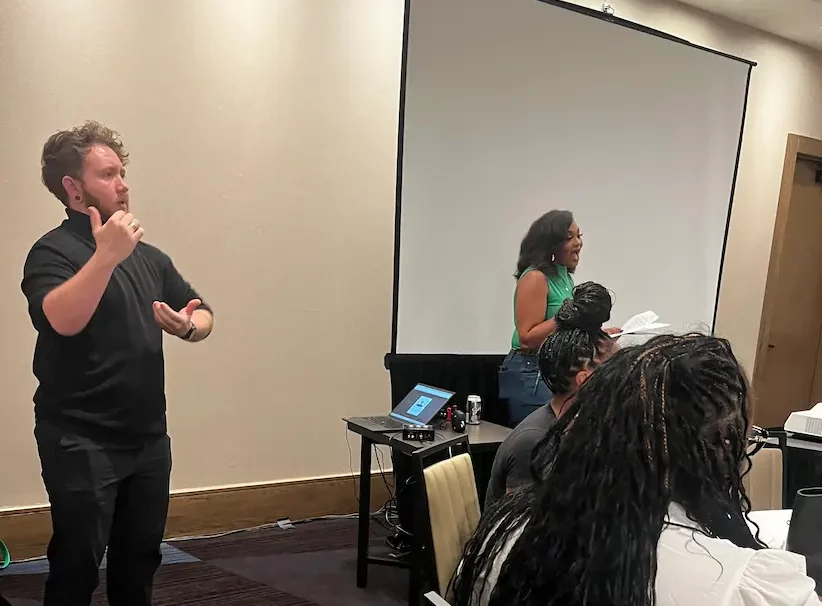 During the event: an ASL interpreter signs at the front of a workshop room while a presenter speaks beside the screen and seated attendees watch.