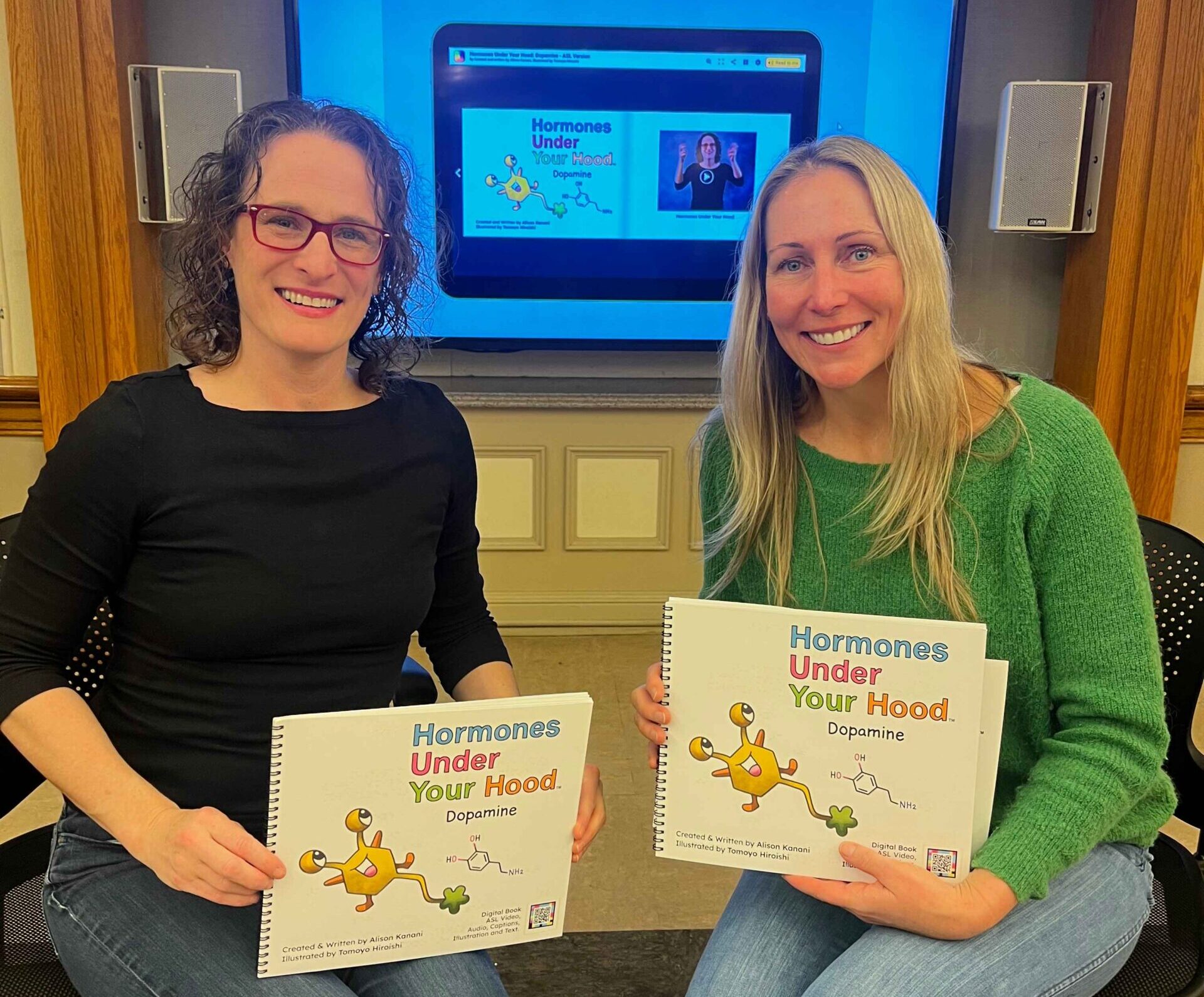 Two people sit side by side holding spiral-bound copies of “Hormones Under Your Hood: Dopamine”, with the digital edition open on the screen behind them showing the book page beside its ASL video panel.