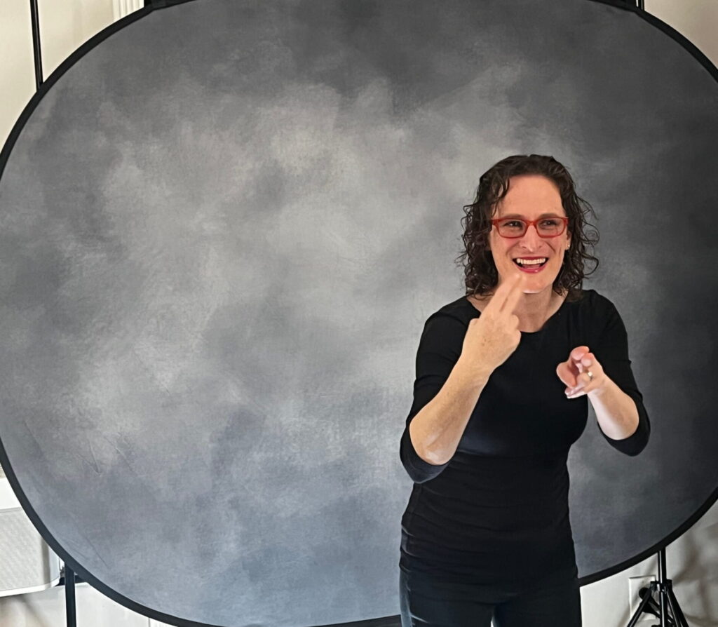 A Deaf interpreter in black stands in front of a grey studio backdrop, smiling as she signs to camera between takes.