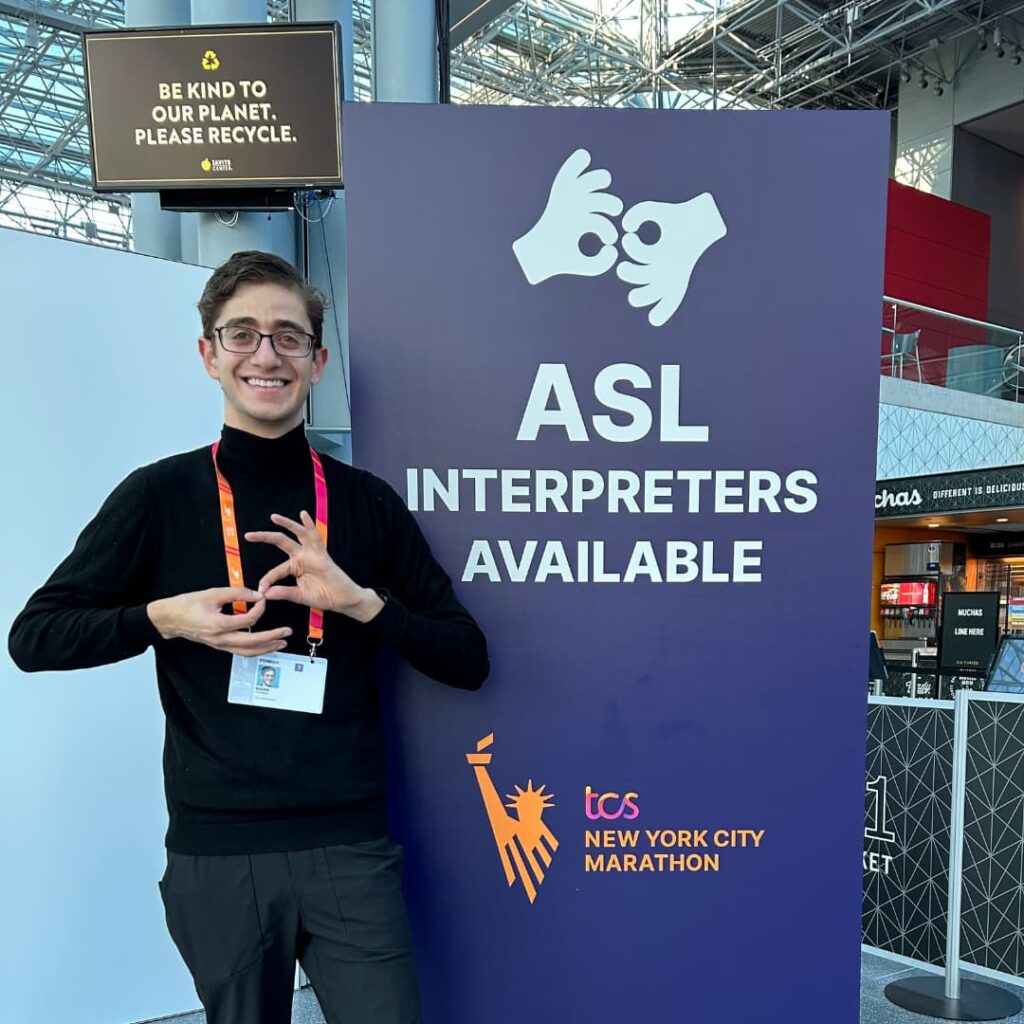 An ICS interpreter smiles and signs beside a tall purple TCS New York City Marathon sign reading 'ASL Interpreters Available' above a sign language handshape icon, inside the marathon expo hall.