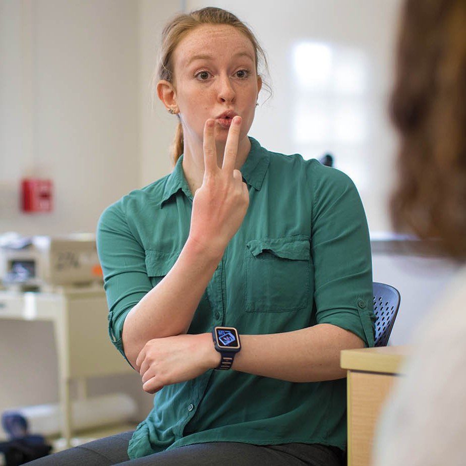 A woman in a green shirt signs during a seated one-on-one conversation in an office.