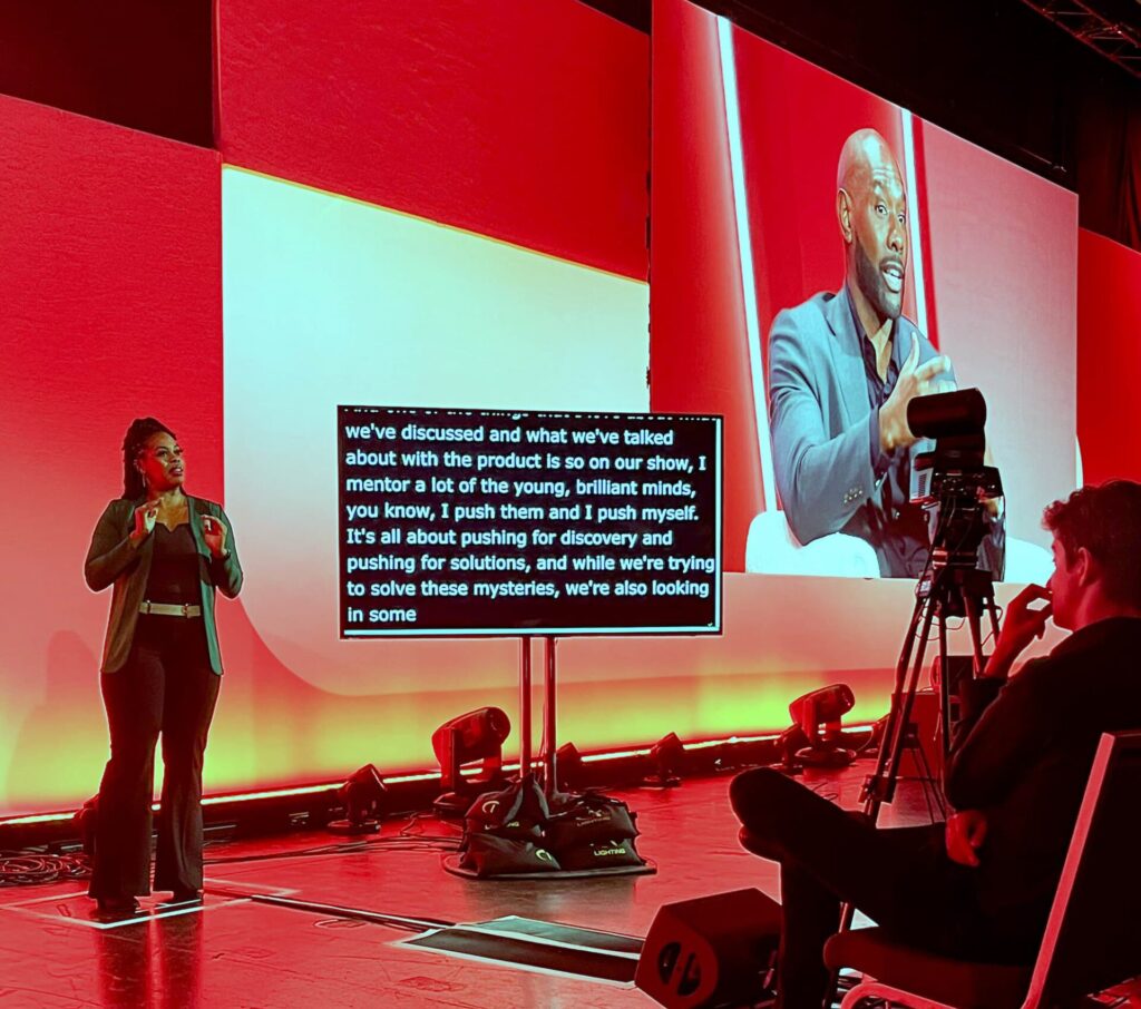 An ASL interpreter signs on the main stage floor beside a live CART caption monitor, with the speaker she is interpreting shown on the large red screen behind her and a camera operator filming from the right.