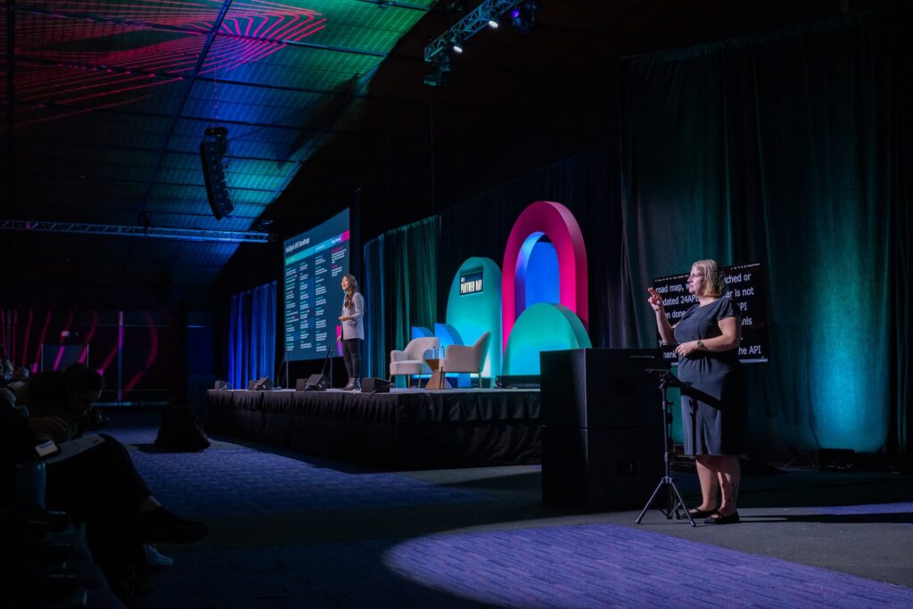 An ASL interpreter signs from a lit spot at the front of a darkened Partner Day room, with the presenter on stage to the left and a CART caption screen glowing behind the interpreter.