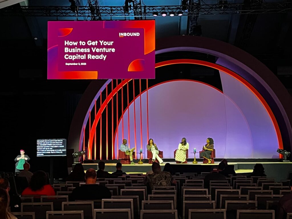 A wide view of the INBOUND stage during the session 'How to Get Your Business Venture Capital Ready', with four panelists seated under the arch, an ASL interpreter at the far left of the room, and a CART caption screen beside her.