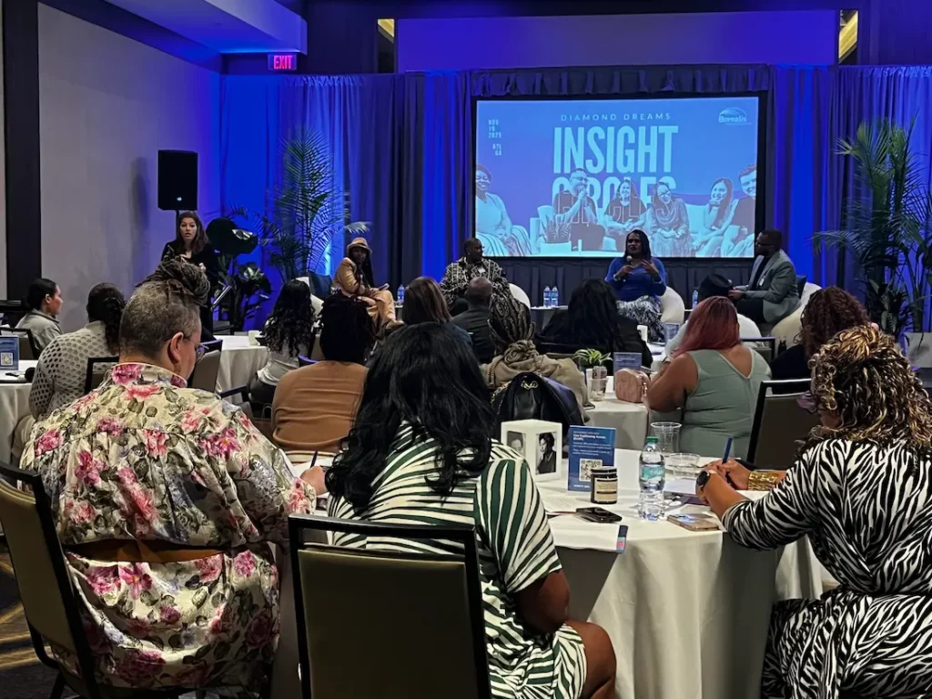 A full workshop room during the Insight Circles session, with panelists seated on stage, an ASL interpreter standing at the left, and attendees at round tables that hold captioning QR code cards.