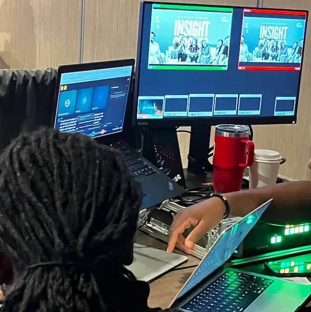 Behind the scenes: a technician works between a laptop and a monitor showing two preview panes of the Insight Circles program feed at the venue tech table.