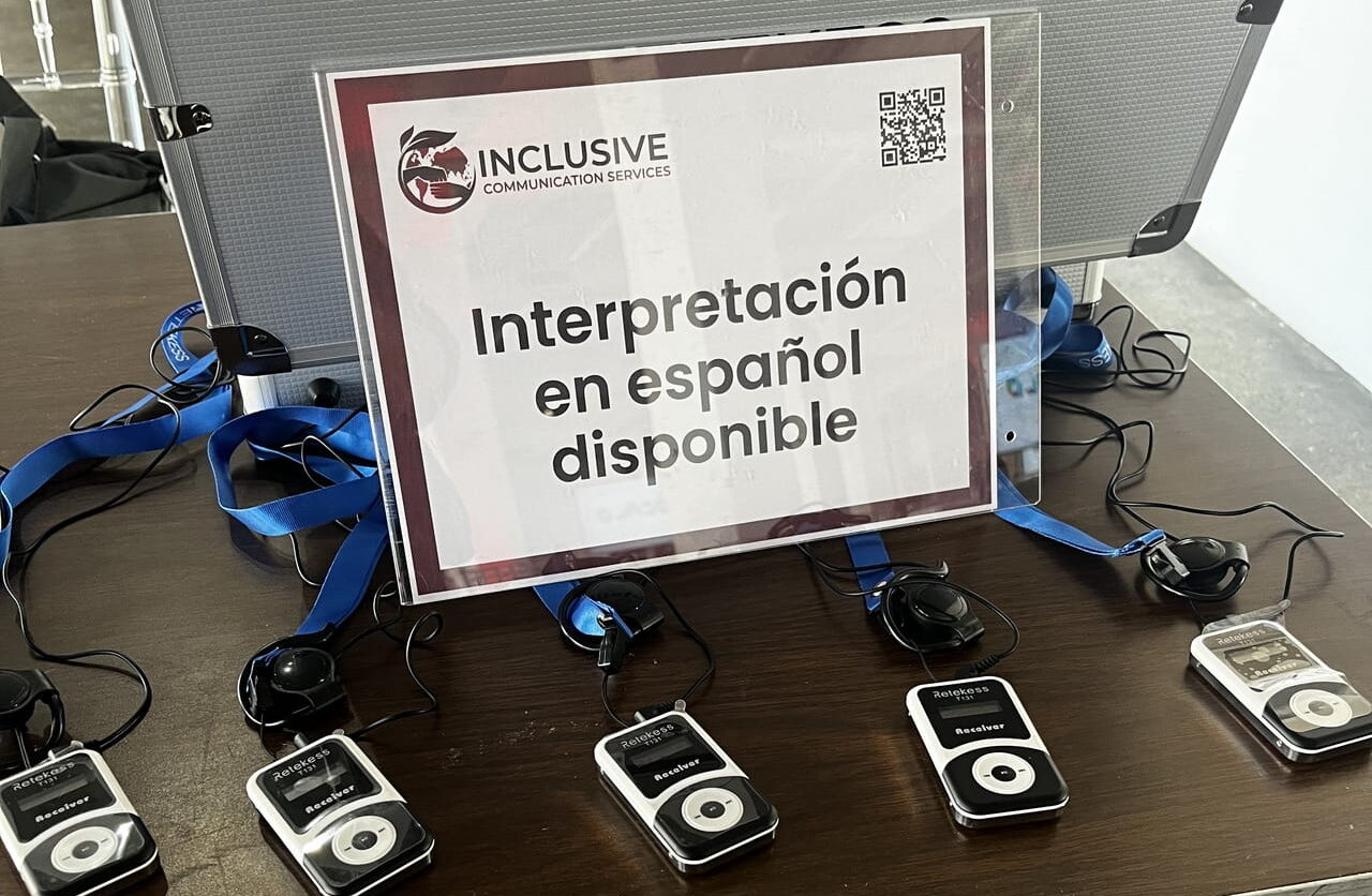 The headset table before doors open: an ICS sign reading 'Interpretación en español disponible' propped against an open flight case, with Retekess receivers and blue lanyards laid out in front of it.