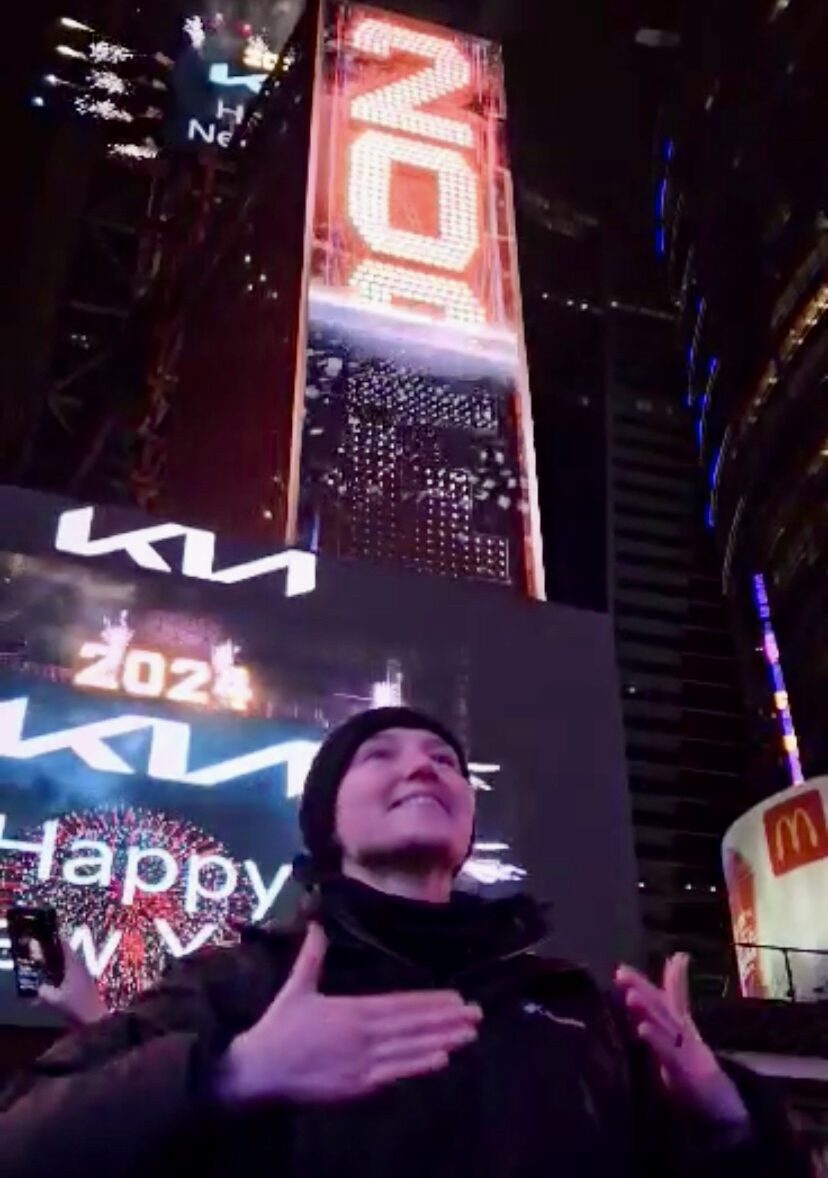 An ASL interpreter in a black parka and beanie signs on the street in Times Square at night, the illuminated 2024 numerals and Kia billboards towering behind her.