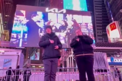 Two ASL interpreters stand side by side on the elevated platform in Times Square, signing in front of the large Kia screen showing the stage performance behind them.