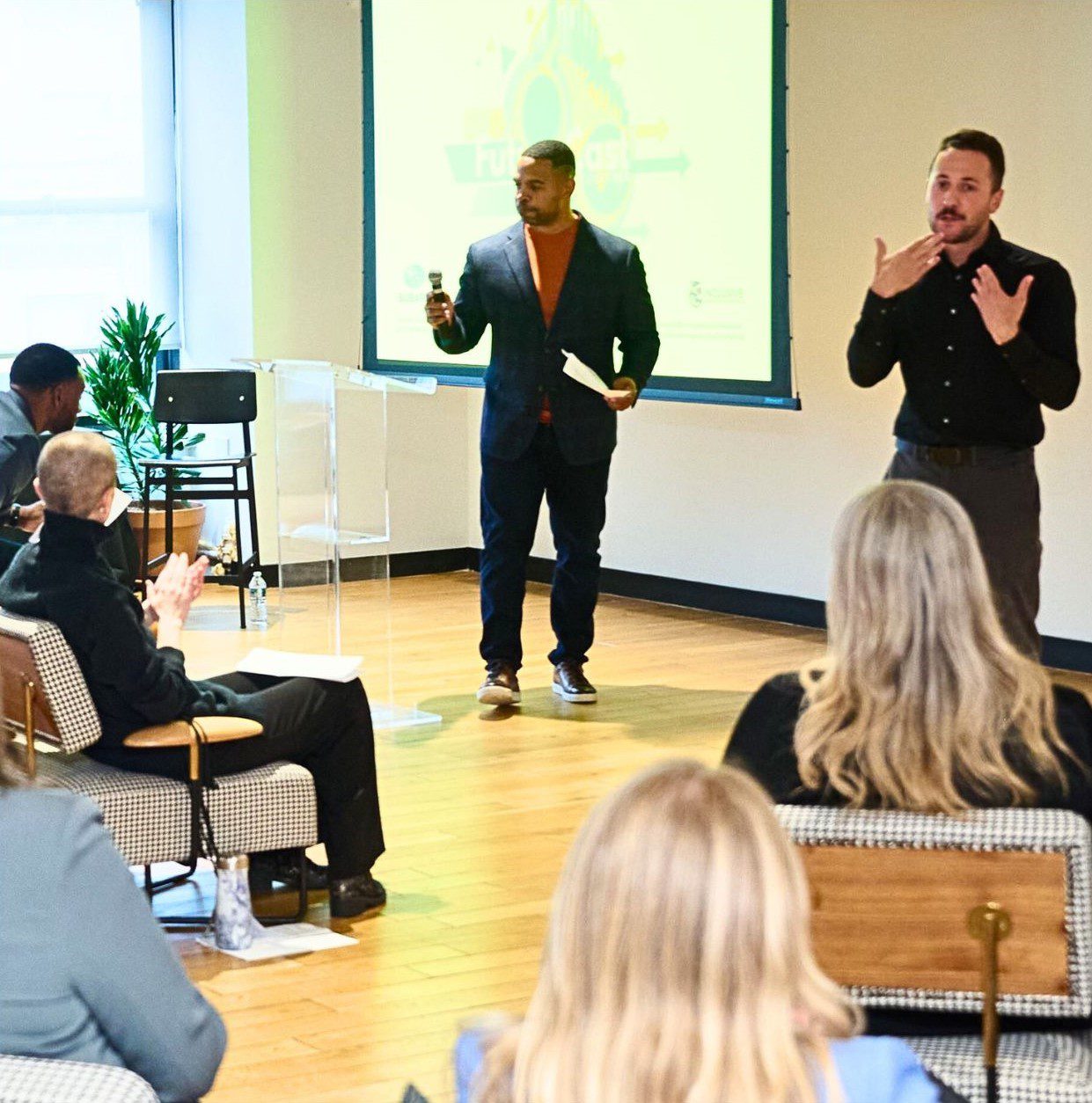 A speaker presenting in a meeting room while an interpreter signs beside him to a seated audience