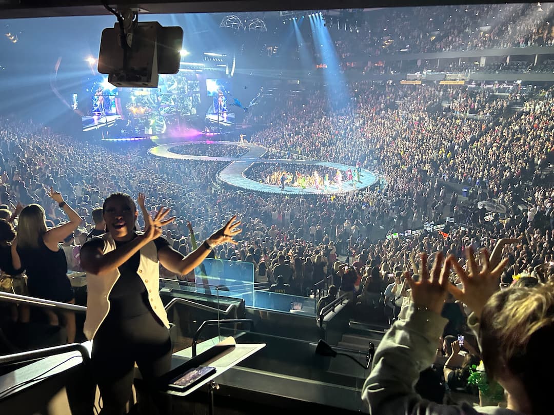 An interpreter signing on a platform beside the stage of a packed arena concert, hands raised throughout the crowd