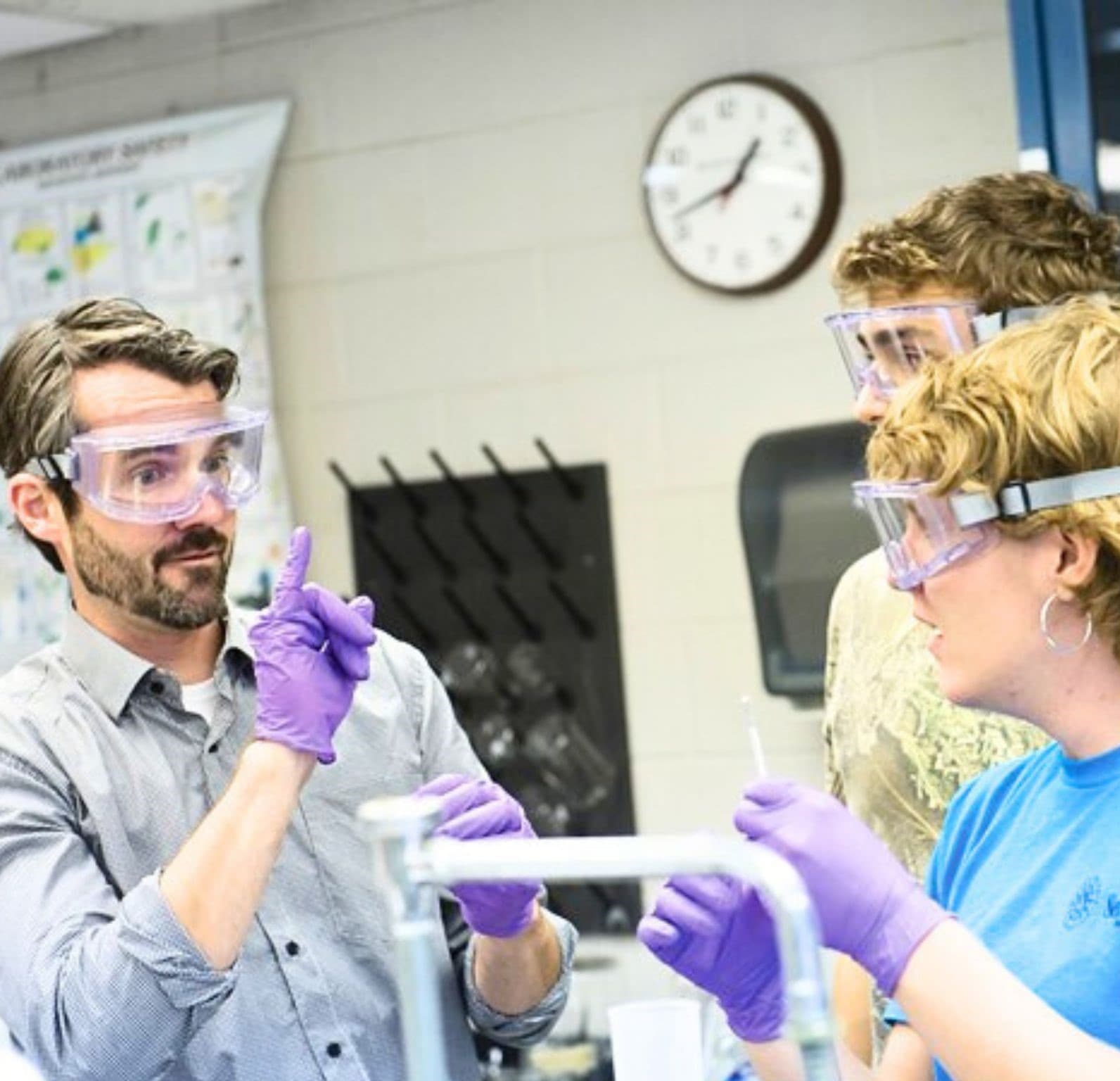 Two people in safety goggles signing to each other across a laboratory bench while a colleague looks on