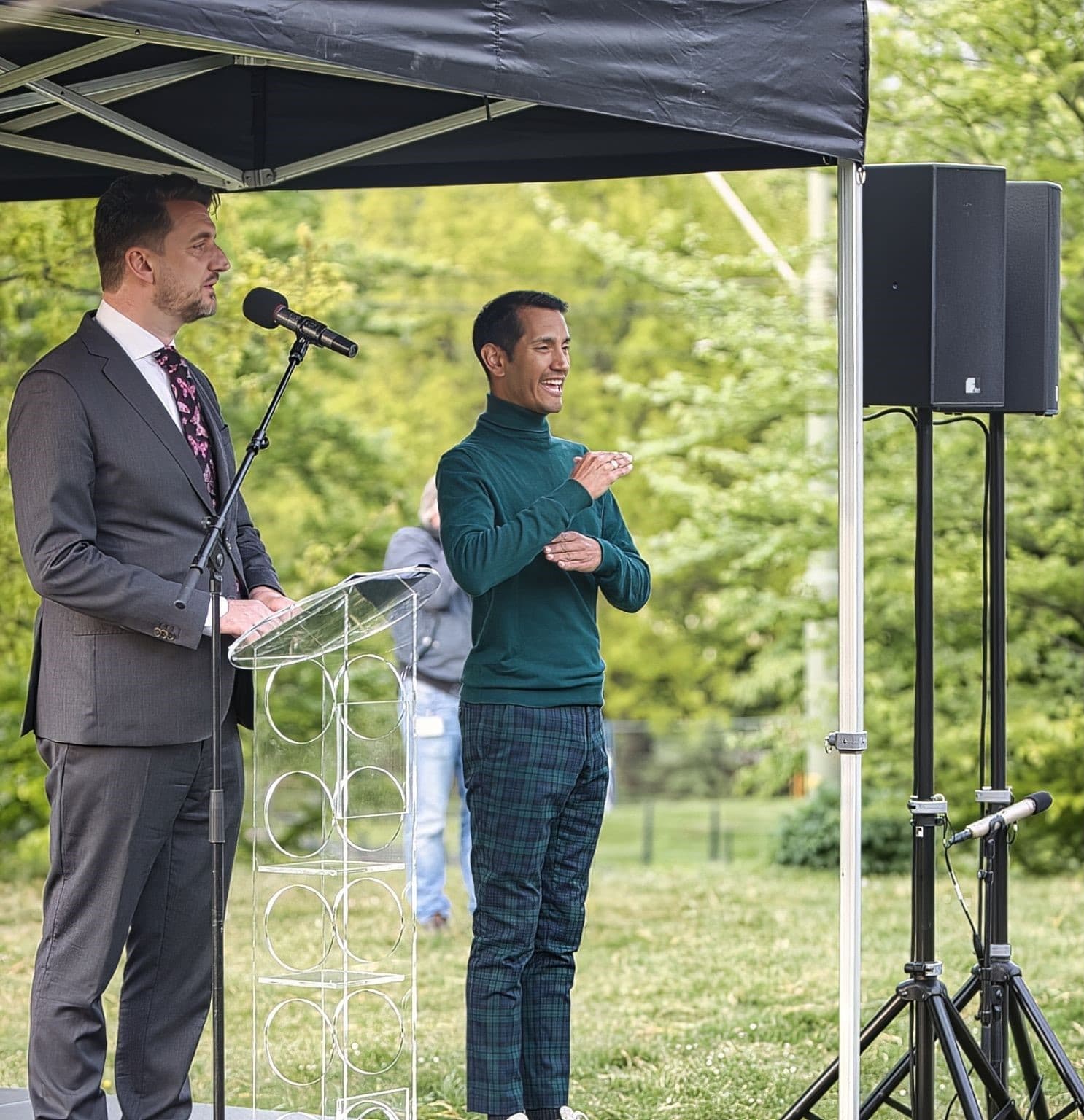 A speaker at an outdoor podium with an interpreter signing beside him under a canopy