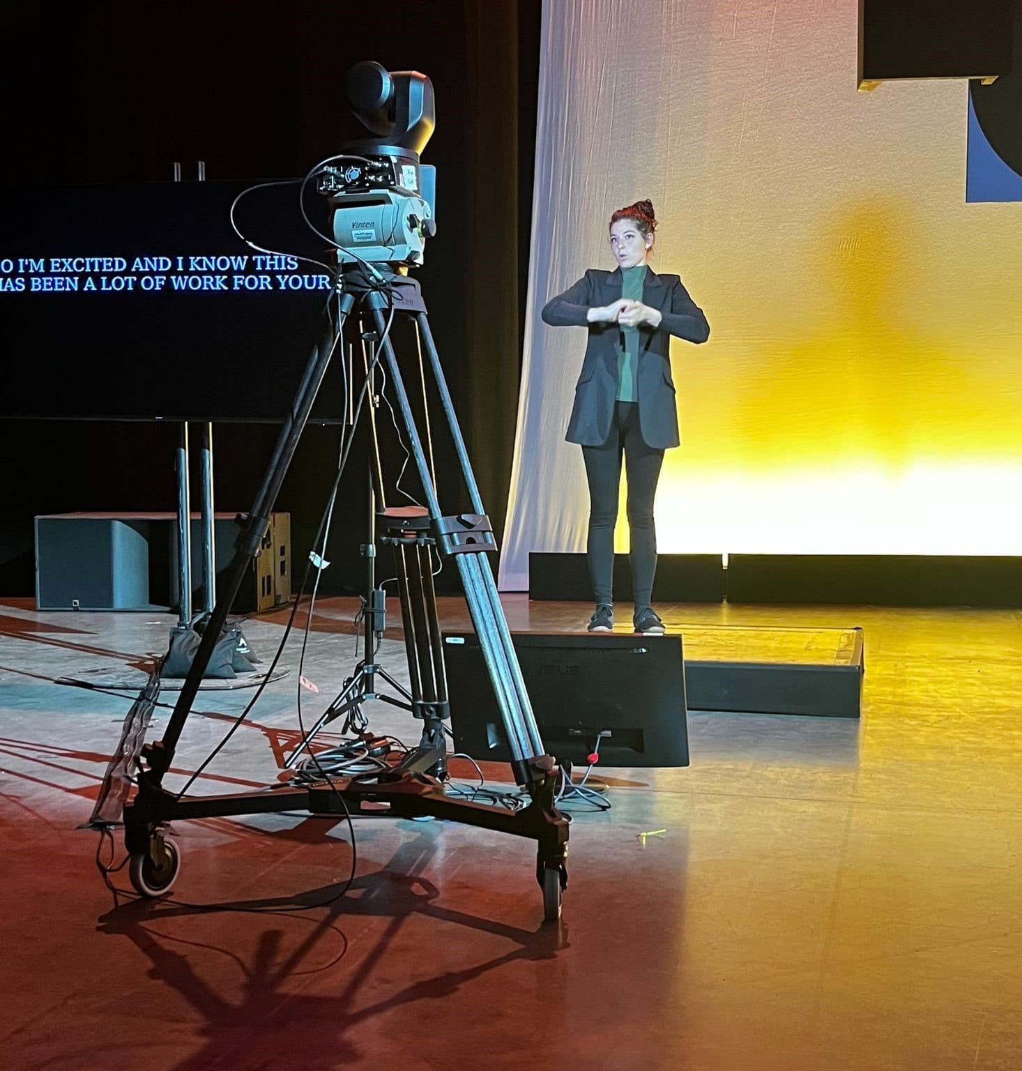 A broadcast camera on a tripod trained on an interpreter signing on a lit studio stage
