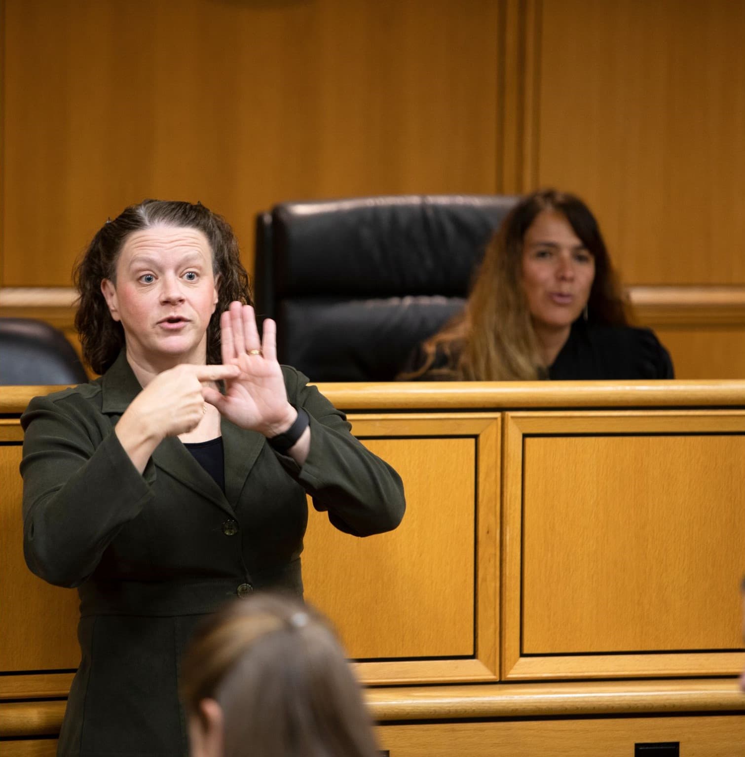 An interpreter signs beside the judge's bench in a wood-panelled courtroom while a court reporter looks on.